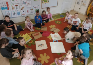 Przedszkolaki siedzą w kole na dywanie, w środku leżą torby na zakupy.