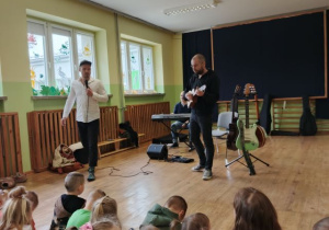 Dzieci siedzą na dywanie i słuchają utworu granego przez muzyka na ukulele.