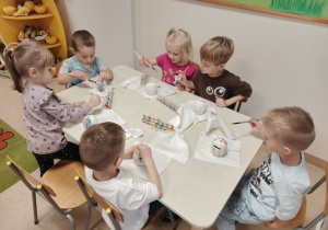 Dzieci siedzą przy stoliku i malują farabami dynie - skarbonki.