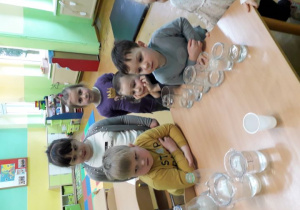 Przedszkolaki przy stoliku z wykorzystaniem szklanych naczyń dokonują pomiaru objętości cieczy w szklanych naczyniach.