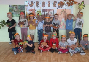 Przedszkolaki w jesiennych opaskach na głowach, trzymające kolorowe listki i witające jesień