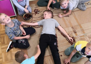 Chłopiec leżący na dywanie, dookoła inni chłopcy układający jego sylwetę z kasztanów