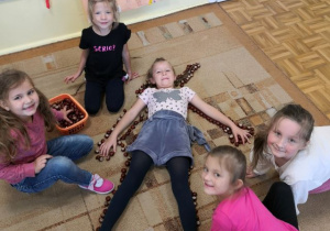 Dziewczynka leżąca na dywanie, dookoła inne dziewczynki układające jej sylwetę z kasztanów