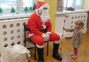 Mikołaj siedzący na krześle i stojaca obok dziewczynka trzymająca laskę Mikołaja
