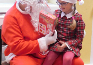 Mikołaj siedzący na krześle trzymający na kolanach dziewczynkę z prezentem
