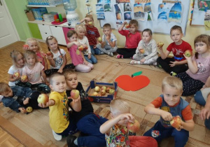 Przedszkolaki siedzące w kole na dywanie, trzymające jabłka