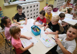 Przedszkolaki siedzące przy stoliku i kolorujące obrazek tematyczny