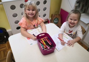 Dziewczynki siedzące przy stoliku i kolorujące obrazek tematyczny