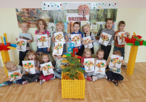 Grupa 3,4,5-latków na tle gazetki tematycznej prezentująca prace plastyczne jesiennych drzew, dookoła stojące drzewa wykonane z kloców.