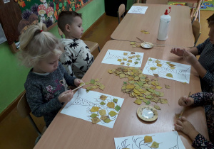 Dzieci przy stolikach nalepiają liście na narysowane drzewo.
