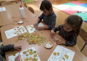 Dzieci przy stolikach nalepiają liście na narysowane drzewo.
