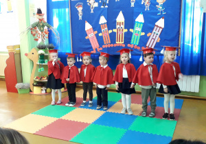 Dzieci w uroczystym stroju (czerwonym birecie i pelerynie) na tle dekoracji PASOWANIE.