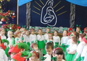 Dziewczynki przebenae za choineczki a chłopcy za elfy ,ustwaione na tle światecznej dekoracji śpiwające pastorałkę