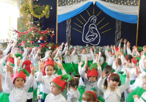 Dzieci z unieśnionymi rękami na tle światecznej dekoracji śpiewające kolędę "Chwała na wysokości"