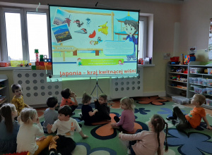 Dzieci siedzą na dywanie i oglądają prezentację multimedialną na dużym ekranie