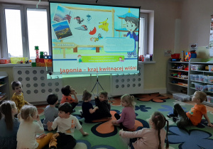 Dzieci z grupy 3,4,5 latków siedzą na dywanie i oglądają prezentację multimedialną na dużym ekranie