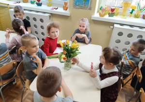 Dzieci siedzące przy stoliku podczas słodkiego poczęstunku. Na stole stoi bukiet tulipanów.