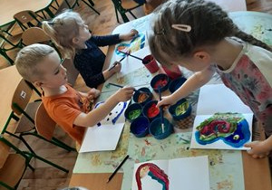 Dzieci z grupy 3,4,5 latków siedzą przy stolikach i malują farbami skrzydła motyla na kartonie.
