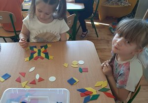 Dziewczynki siedząc przy stoliku układają kształty motyla z kolorowych klocków w kształcie figur geometrycznych.