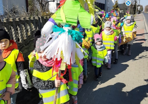 Wiosenny pochód przedszkolaków. dzieci idą chodnikiem w grupie, dziecko z przodu trzyma marzannę.