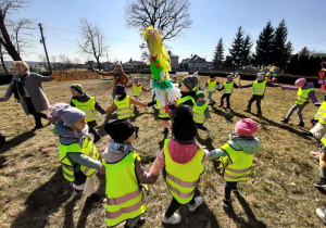 Na dworze. Duża grupa dzieci tańczy w kole z marzanną na placu porośniętym trawą.