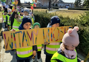 Dzieci spacerujące po chodniku w parach. Przedszkolaki z przodu trzymają transparent z napisem witamy wiosnę.