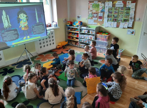 Grupa dzieci siedząca przed rozkładanym ekranem, oglądająca prezentację multimedialną o kosmosie.