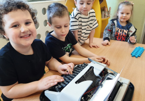Kilkoro dzieci siedzących przy maszynie do pisania. Chłopiec pisze na maszynie.