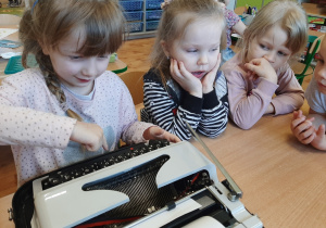 Trzy dziewczynki siedzące przy maszynie do pisania, jedna z nich pisze na maszynie.
