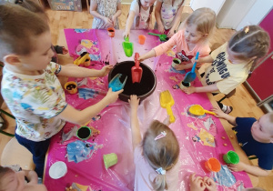 Dzieci z grupy 3,4,5 latków siedzące przy stole, wsypujące łopatkami ziemię do kolorowych doniczek.
