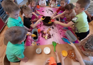Grupa dzieci 5, 6 letnich siedząca przy dużym stole. Dzieci wsypują łopatkami ziemię do kolorowych doniczek, sadzą cebulę.