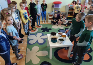 Przedszkolaki z grupy 5,6 latków oglądają naczynia z naturalnymi barwnikami, których opowiadają ich dwaj koledzy stojący przy stoliku.