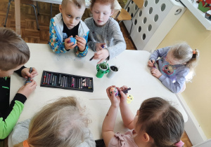 Dzieci 5, 6 letnie siedzące przy stoliku, malujące jajka pastelami. Kolejny stolik.