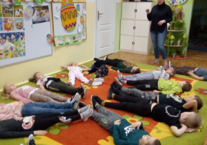 Dzieci z grupy 5,6 latków leżą na plecach na dywanie i wykonują ćwiczenie proponowane przez panią logopedę