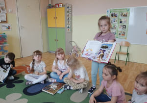 Dzieci siedzące na dywanie, dziewczynka prezentuje im swoją ulubioną książkę z bajkami.