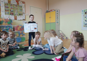 Dzieci siedzące na dywanie, chłopiec prezentuje im swoją ulubioną książkę - atlas dla dzieci.