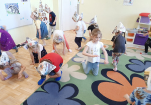 Dzieci w gazetowych czapeczkach naśladują ruchem przedzieranie się przez leśny gąszcz.