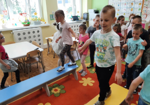 Dzieci stoją w dwóch rzędach, przechodzą po ławeczkach gimnastycznych.