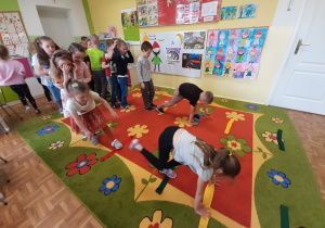 Dzieci ustawione w dwóch rzędach przechodzące na czworakach po drabince ułożonej na podłodze z szarf gimnastycznych.