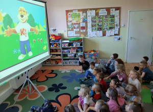 Dzieci siedzące na dywanie oglądają wyświetlaną na dużym ekranie animację o misiu Kubusiu.