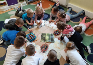 Dzieci skupione w kole na dywanie wykonują elementy do plakatu na temat lasu.