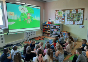 Dzieci siedzące na dywanie oglądają wyświetlaną na dużym ekranie animację o misiu Kubusiu.