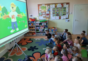 Dzieci siedzące na dywanie oglądają wyświetlaną na dużym ekranie animację o misiu Kubusiu, inne ujęcie..