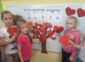 Dziewczynki z grupy 5,6 latków prezentują na tle drzewka dobrych uczynków czerwone szablony serc.