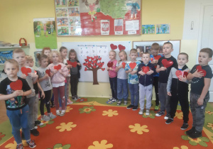 Dzieci z grupy 5,6 latków stoją przy zawiedszonym na tablicy papierowym drzewku szczęścia, w rękach trzymają szablony czerwonych serc.