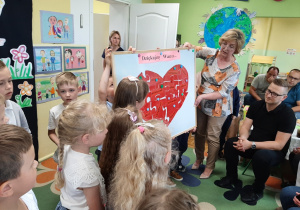 Dzieci wraz z nauczycielem prezentują rodzicom tablicę magnetyczną, na której przyczepione są puzzle złożone w kształt serca dla rodziców.