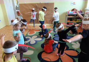 Grupa dzieci 3,4,5 letnich w opaskach z bocianim dziobem uczestniczy w zabawie ruchowej na dywanie.