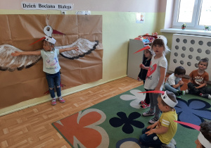 dziecko z grupy 3,4,5 latków w opasce z bocianim dziobem mierzy rozpiętość skrzydeł bociana namalowanego na dużym arkuszu papieru, przyczepionym do ściany. Reszta grupy obserwuje.