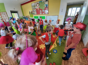 Dzieci w kolorowych strojach tańczą w grupach, w parach. Chłopiec w czerwonym kapeluszu śpiewa piosenkę do mikrofonu.