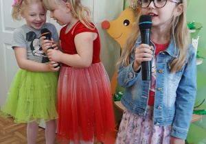 Trzy dziewczynki w kolorowych strojach śpiewają piosenkę do mikrofonów.